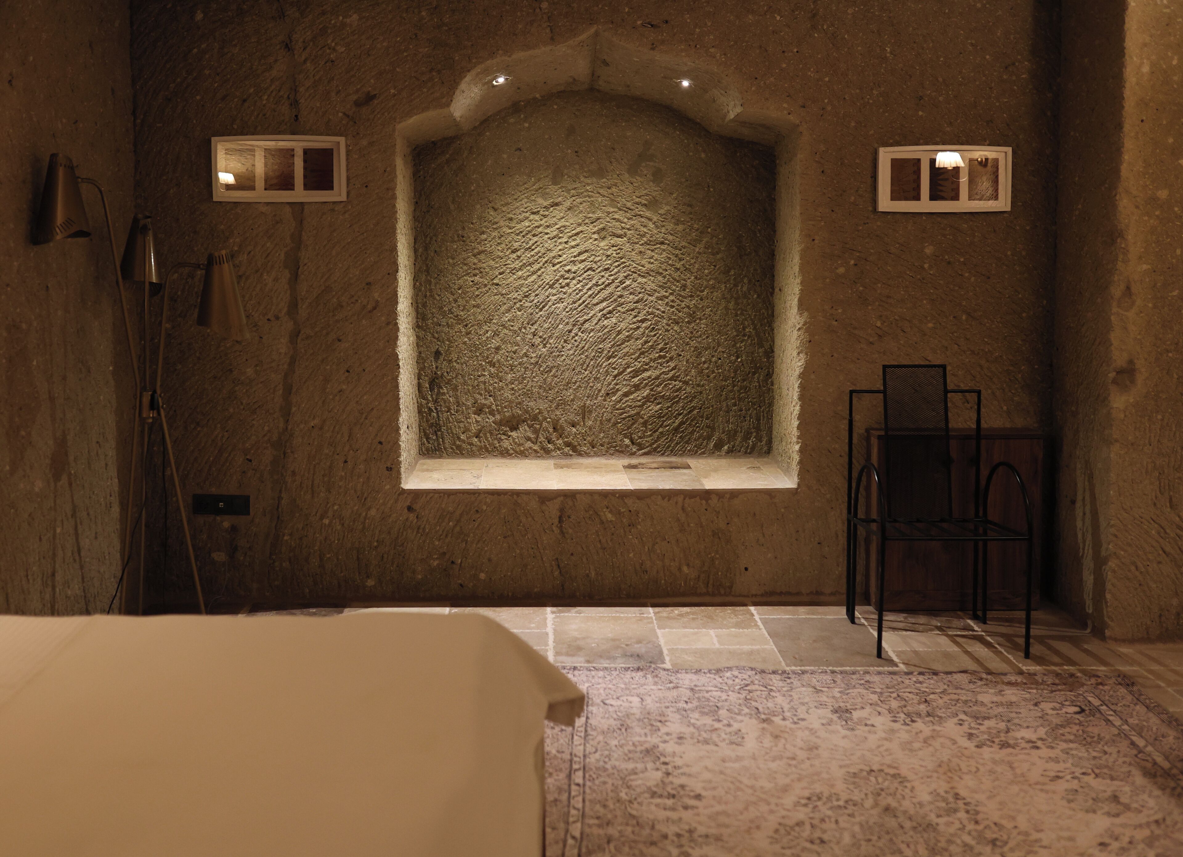 A room with a stone wall, a window with a textured design, a bed, a chair, and a small table.