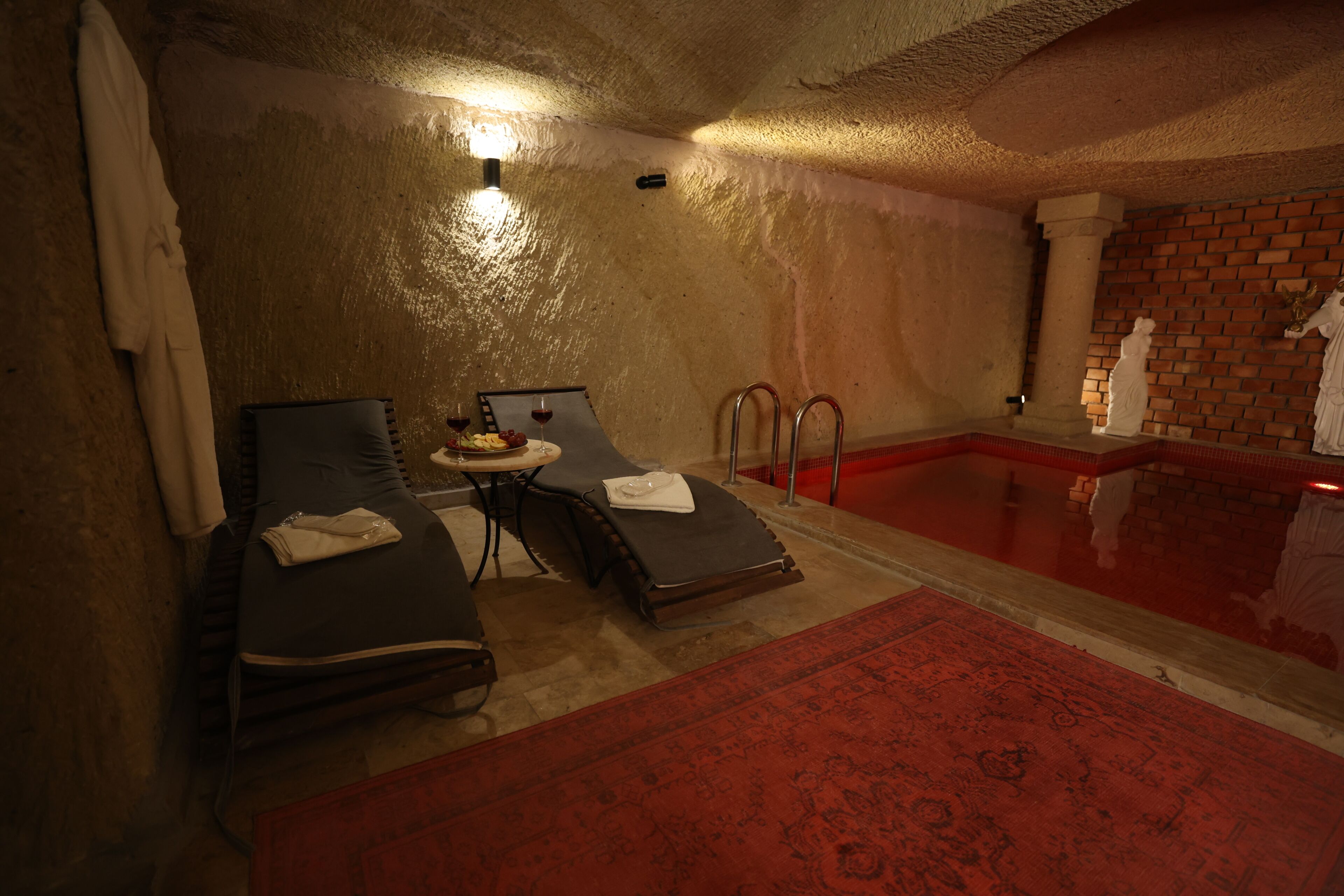 Un cuarto de spa con dos sillones reclinables, una mesita con frutas y vino, un tapete rojo estampado y una piscina con azulejos rojos.