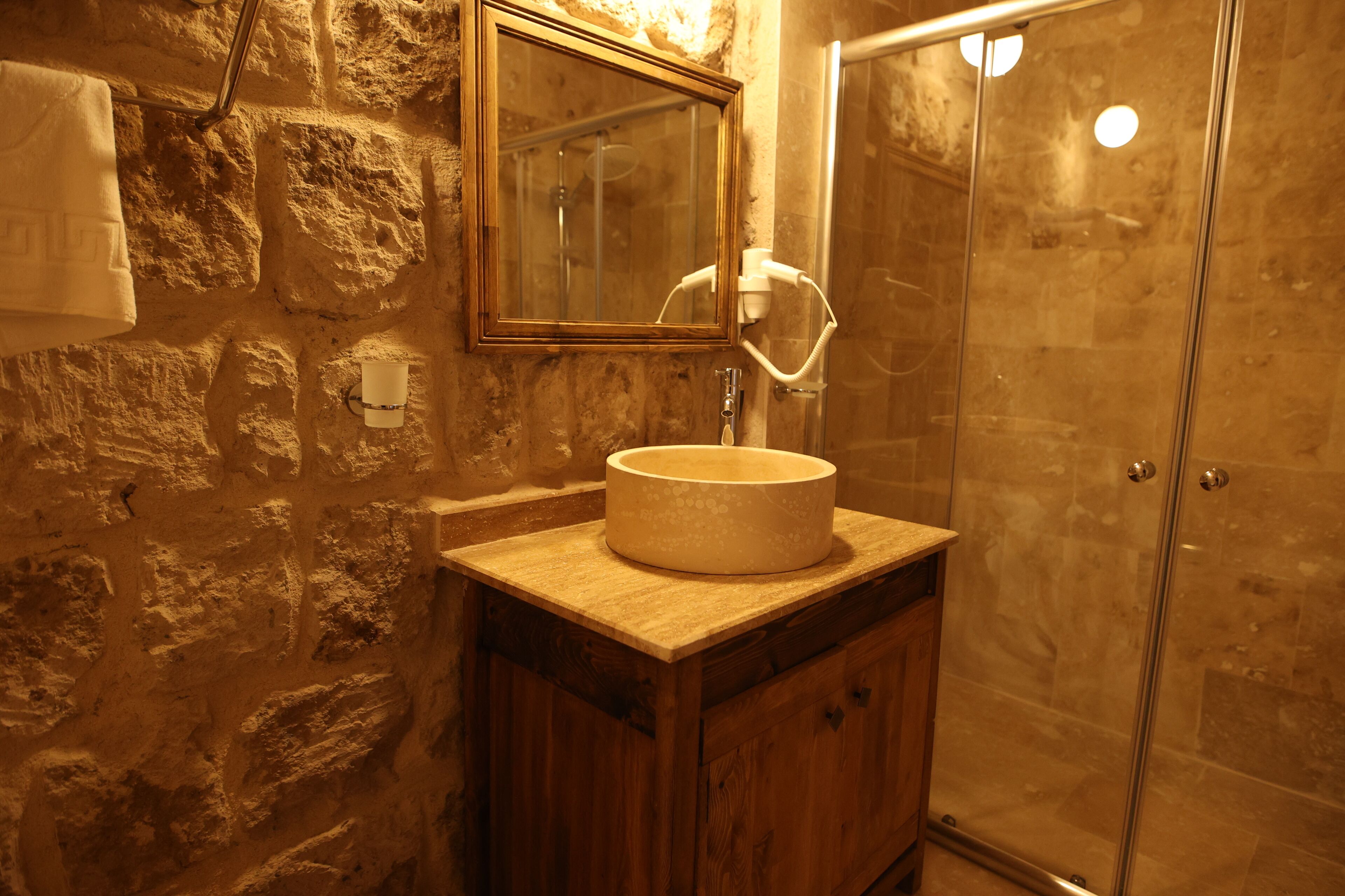 A bathroom with a stone wall, a wooden vanity, a round sink, a mirror, and a glass shower enclosure.