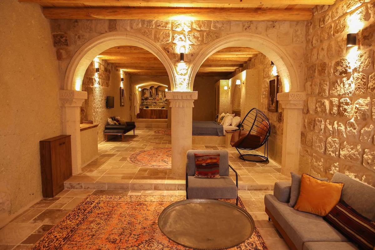 Un hall de hotel con paredes de piedra, una zona de estar central con un sofá y sillones, y una alfombra estampada.