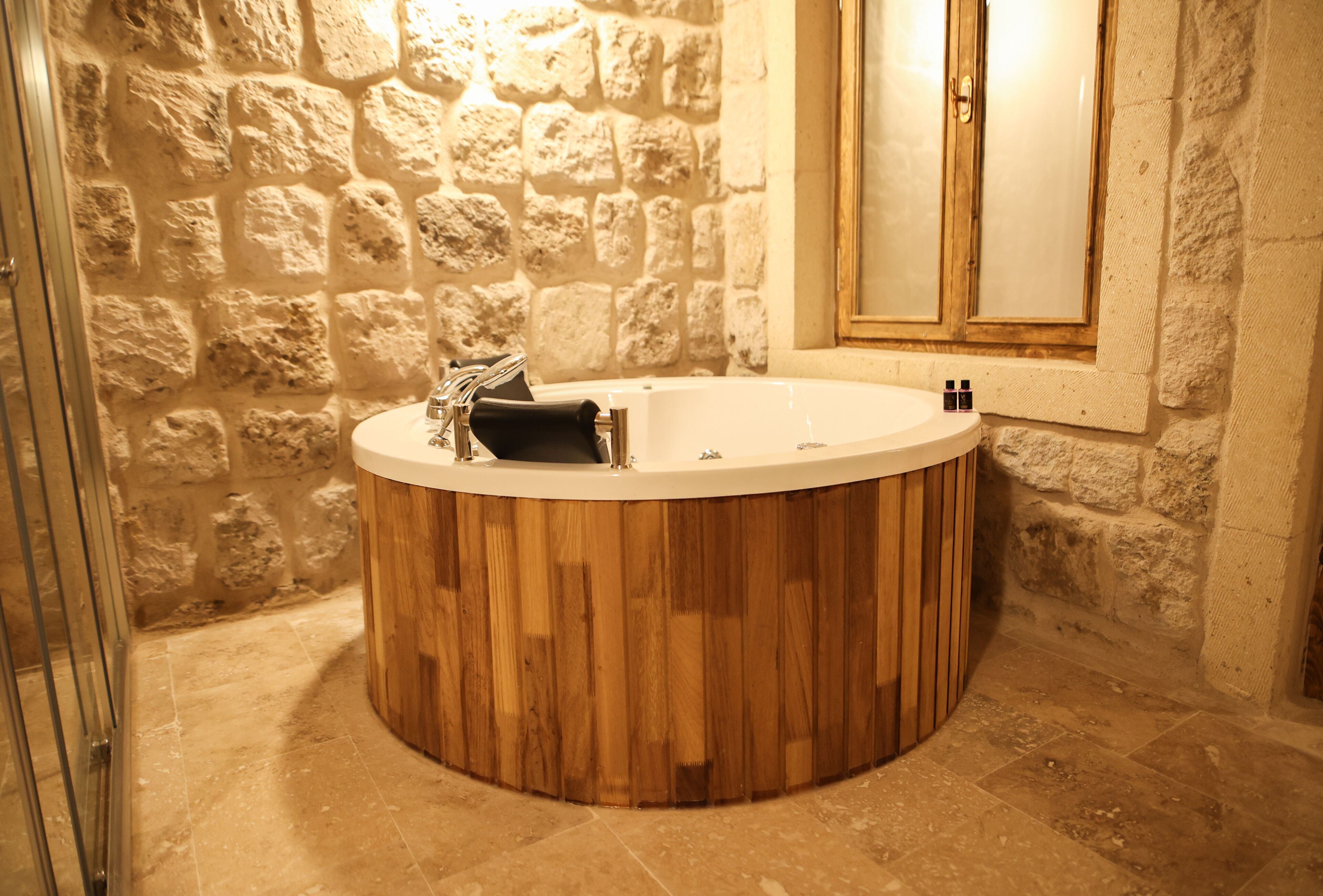 A circular wooden bathtub in a stone-walled bathroom with a glass shower enclosure and a wooden door.
