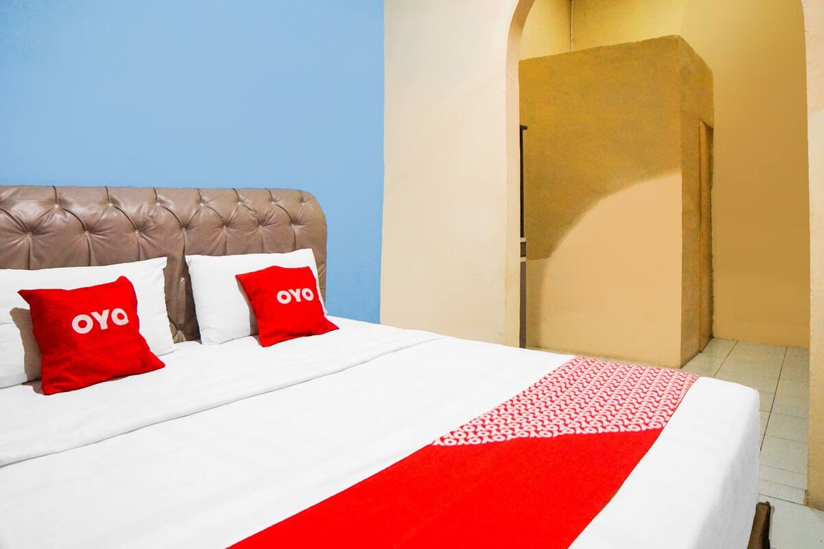 A hotel room with a tufted headboard, two red pillows with "OYO" branding, a white bedspread, and a red and white patterned throw.