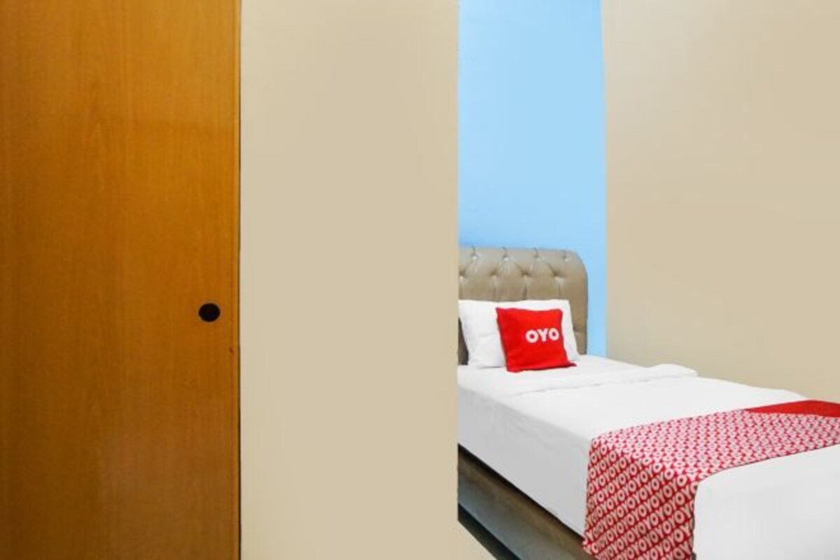 A hotel room with a wooden door, a bed with a white mattress, a red pillow, and a red and white patterned blanket.