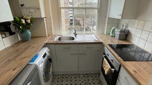 A compact kitchen with a washing machine, a sink, a stove, and a window with a view of buildings. - Beautiful Vintage Style 1BD Apartment - Pimlico (London)