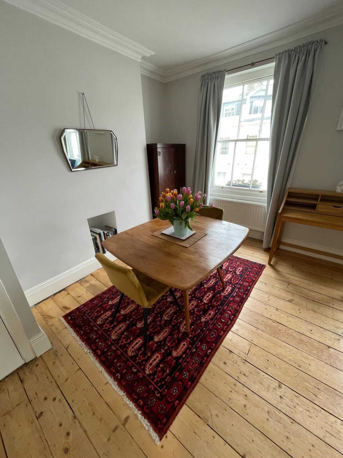 Un comedor con una mesa de madera, sillas, un tapete rojo estampado, un jarrón con flores, un aparador de madera y una ventana con cortinas.