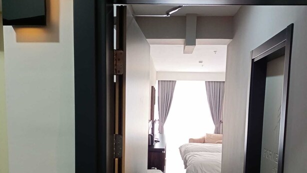 Pasillo de hotel que conduce a un dormitorio con cama, mesita de noche y ventana con cortinas.