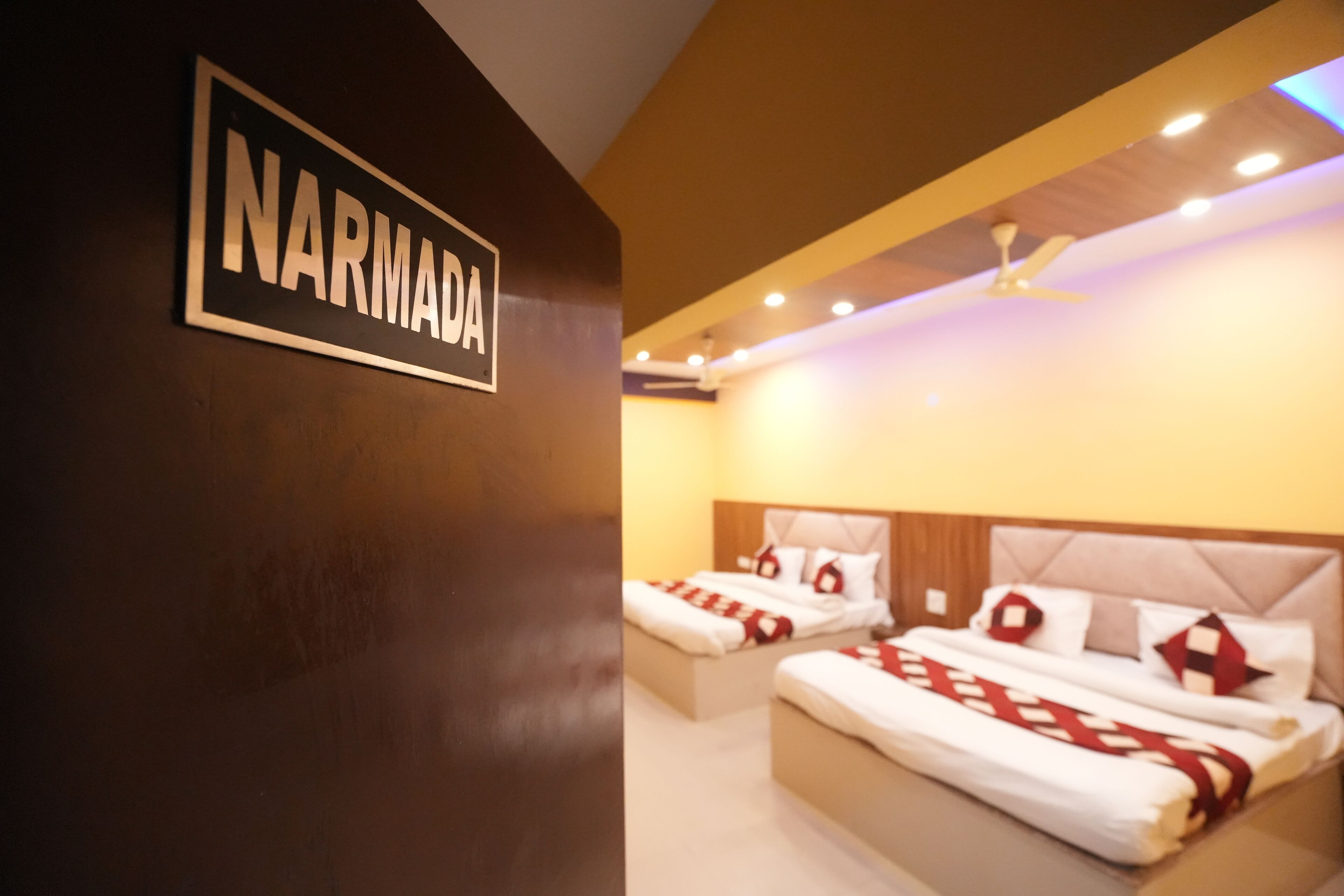 A hotel room with two beds, a ceiling fan, and a sign that reads "NARMADA".