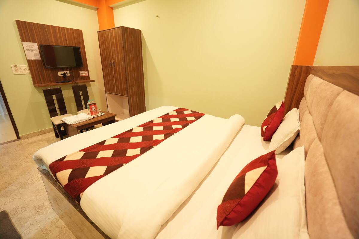 A hotel room with a bed, a TV mounted on the wall, a wooden wardrobe, and a small table with a bottle and papers.