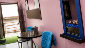 Een kamer met een roze muur, een bed met een blauw hoofdeinde, een raam, een tafel met een stoel en een klein balkon met een bloempot.