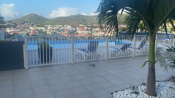 A pool area with a view of a town, a palm tree, and a fence. - Spacious apartment with pool and gorgeous views of Marigot! (Marigot)