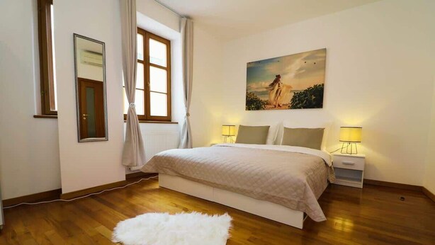Een slaapkamer met een groot bed, een raam met gordijnen, een schilderij aan de muur en een witte vloerkleed.