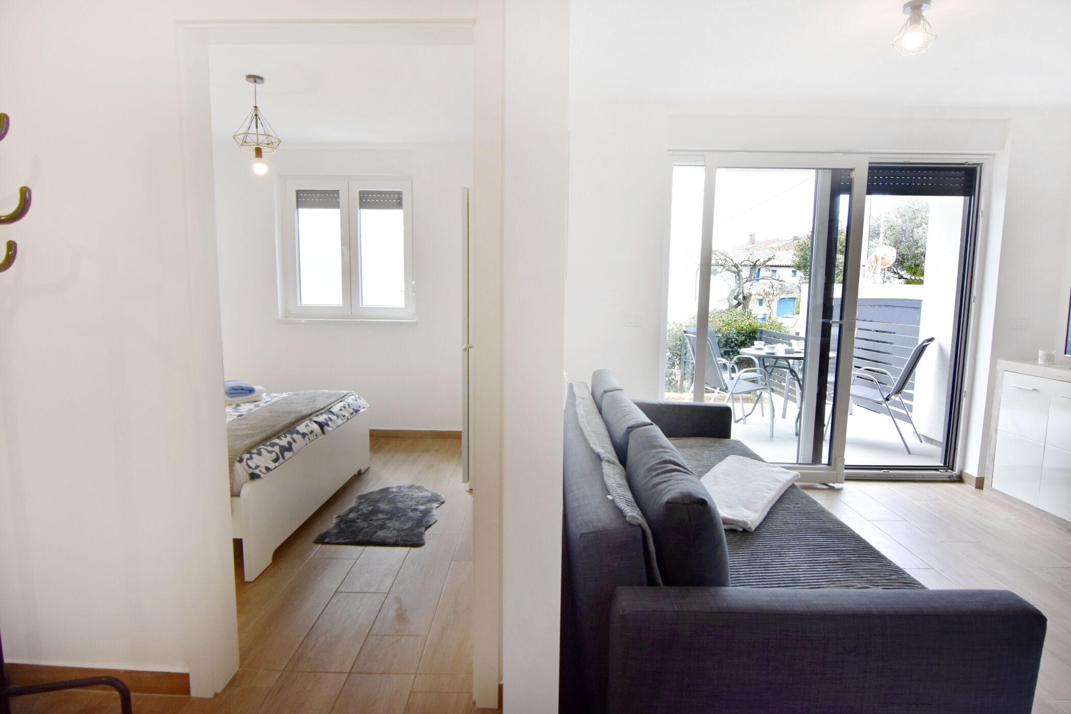 A modern living room with a sofa, a small table, and a window. There is a door leading to a balcony with outdoor furniture.