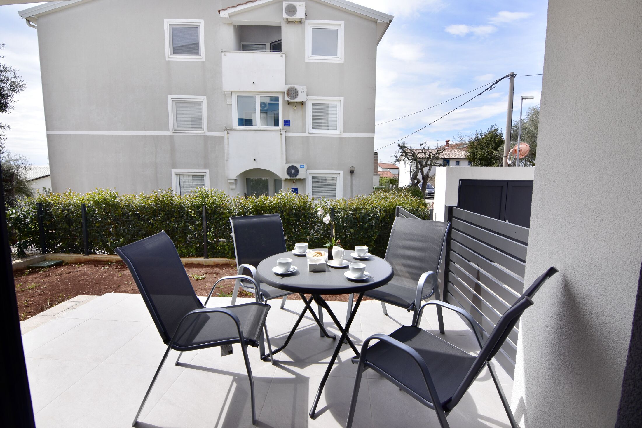 Eine Terrasse im Freien mit einem runden Tisch und vier Stühlen, einem kleinen Tisch mit Tassen und einem Teller sowie einem Gebäude im Hintergrund.