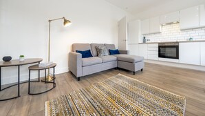 A modern living room with a grey sectional sofa, a small round table, a floor lamp, and a patterned rug. - Stylish split-level 2BR near Queen’s Park – bright, modern & well-connected (London)
