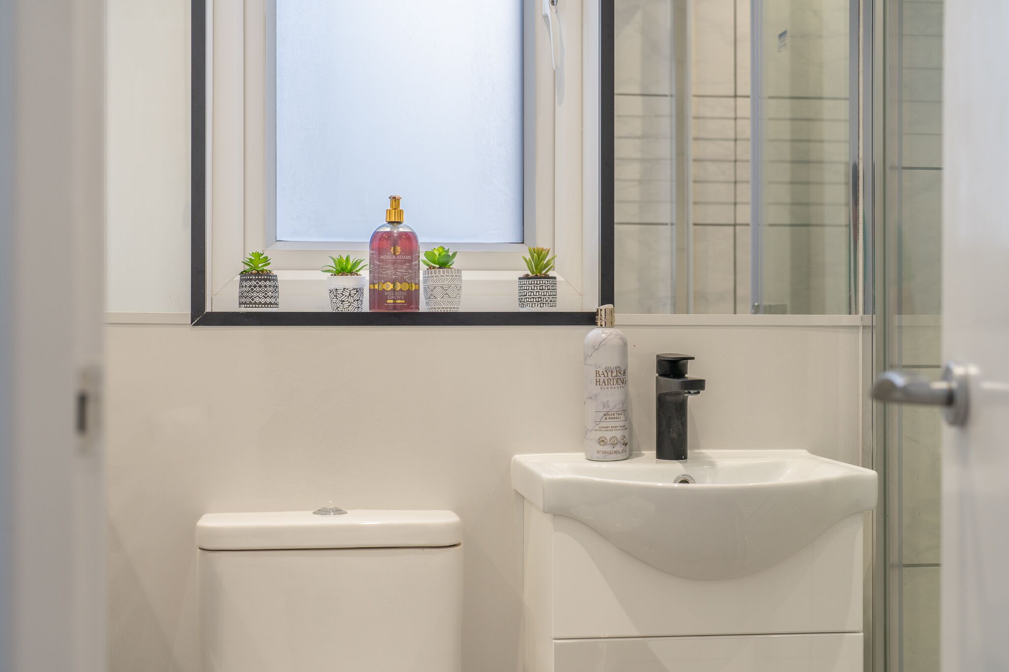 A bathroom with a toilet, sink, and window. There are small potted plants on the windowsill and a bottle of hand soap.