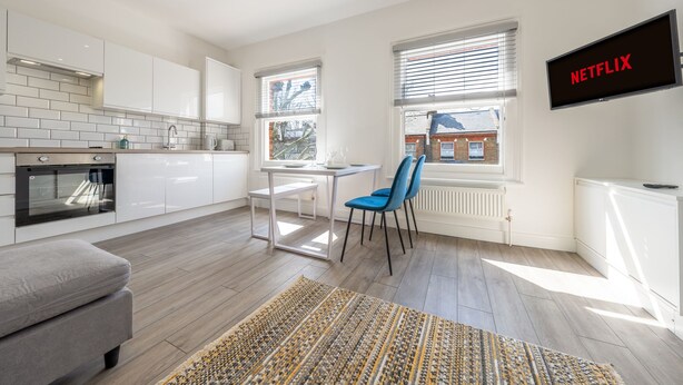 A modern kitchen with white cabinets, a dining table, and chairs. A flat-screen TV mounted on the wall displays "NETFLIX". - Stylish split-level 2BR near Queen’s Park – bright, modern & well-connected (London)