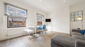 A modern living room with a grey sofa, a dining table with chairs, a TV mounted on the wall, and large windows with blinds. - Stylish split-level 2BR near Queen’s Park – bright, modern & well-connected (London)