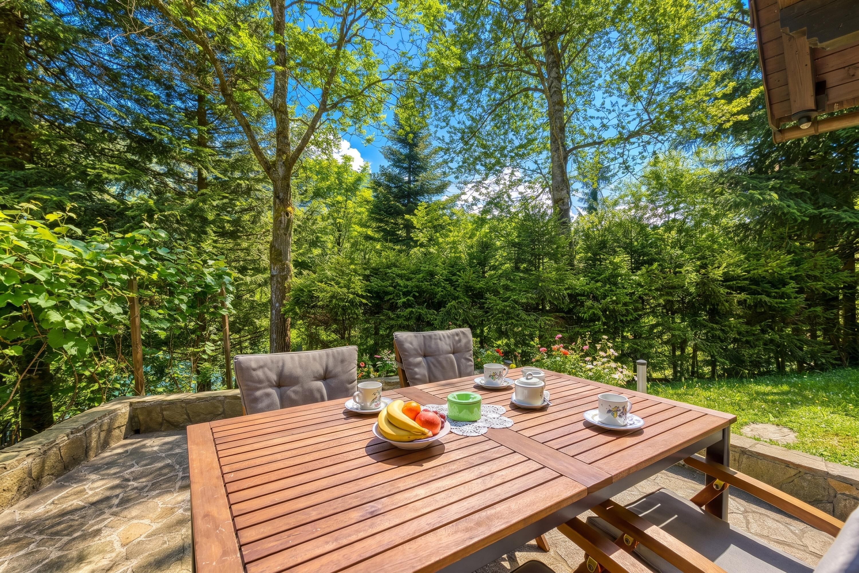 An outdoor wooden table with chairs, surrounded by lush greenery and trees.