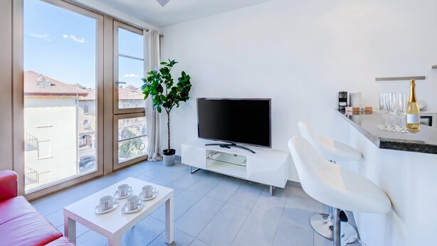 A modern living room with a flat-screen TV, a dining area with a table and chairs, a potted plant, and a balcony with a view of buildings. - Residenza Castello Flat With A/C and Garage (Mendrisio)