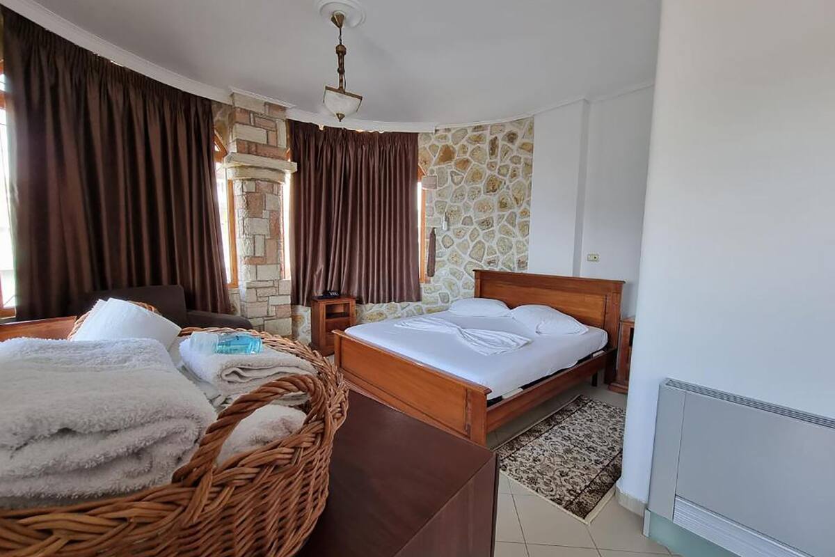 A bedroom with a bed, a wicker basket, a nightstand, a stone wall, and a window with curtains.