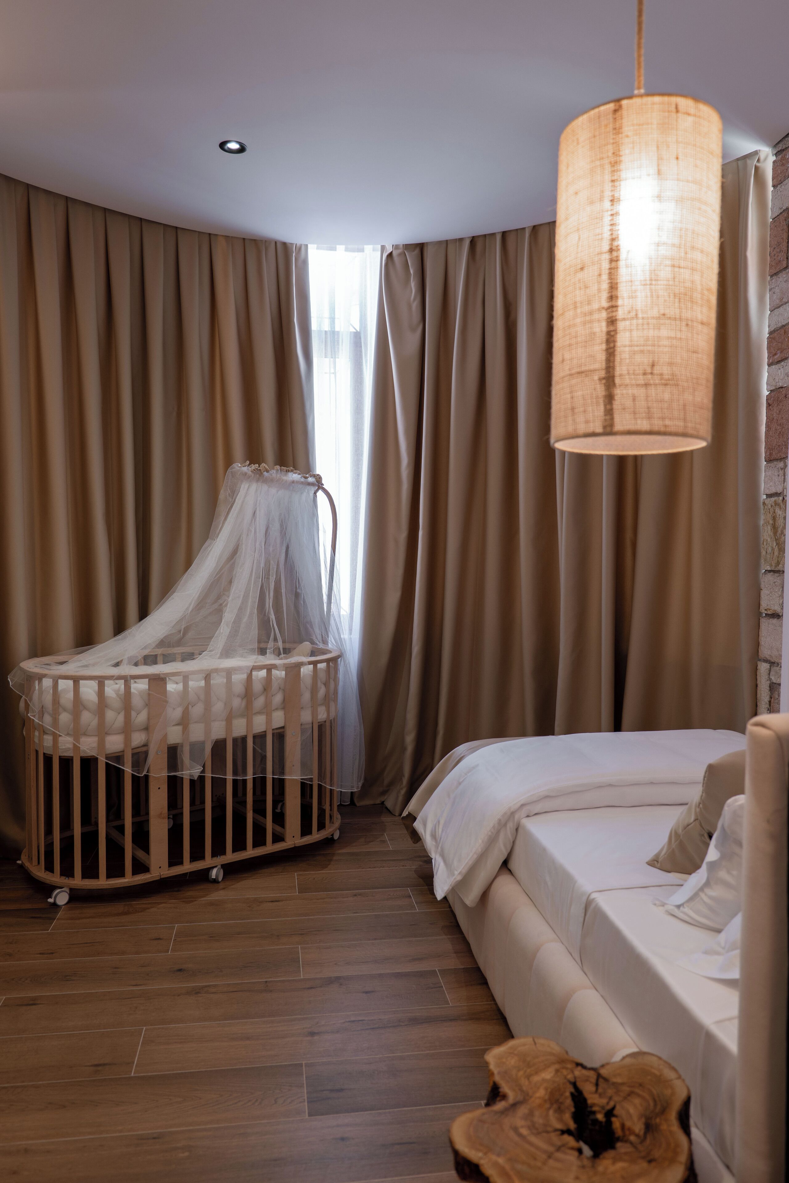 Ein Zimmer mit einem Kinderbett, einem Bett, einer Hängelampe und einem großen Fenster mit Vorhängen.