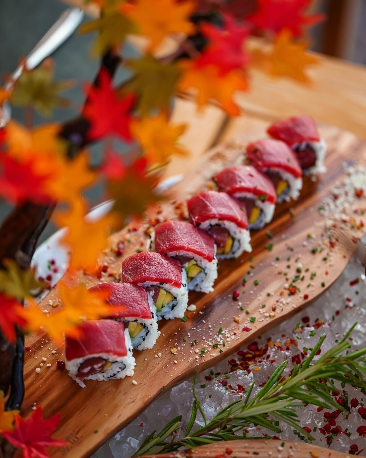 Une planche de service en bois, sur laquelle sont disposés des sushis, décorés d’herbes aromatiques et d’épices, et entourés de feuilles d’automne.