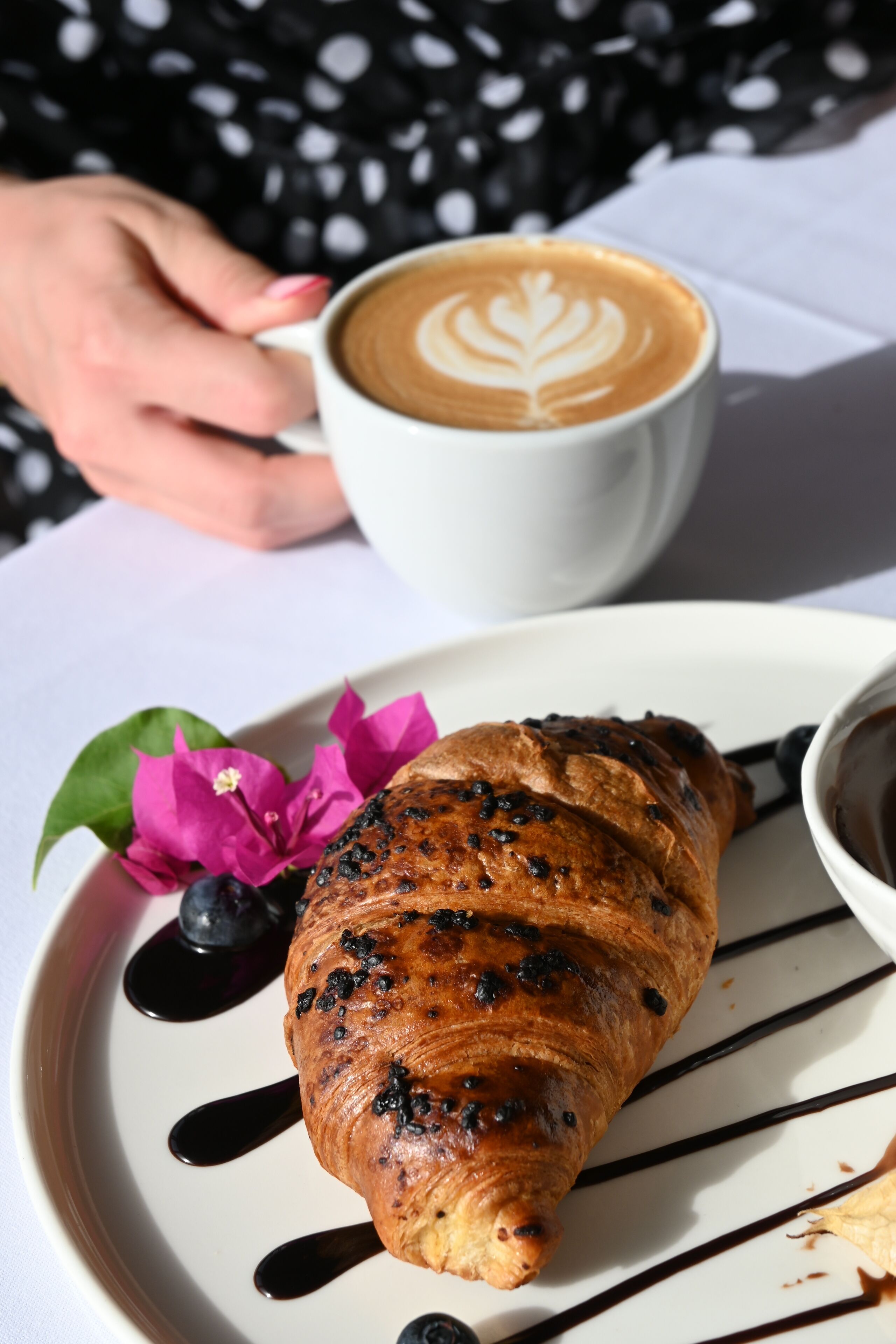 Un croissant con glaseado de chocolate y arándanos sobre un plato, una taza de café y una flor rosa.