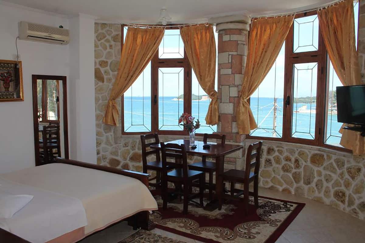 A bedroom with a bed, a dining table with chairs, a TV, and a view of the sea through large windows.