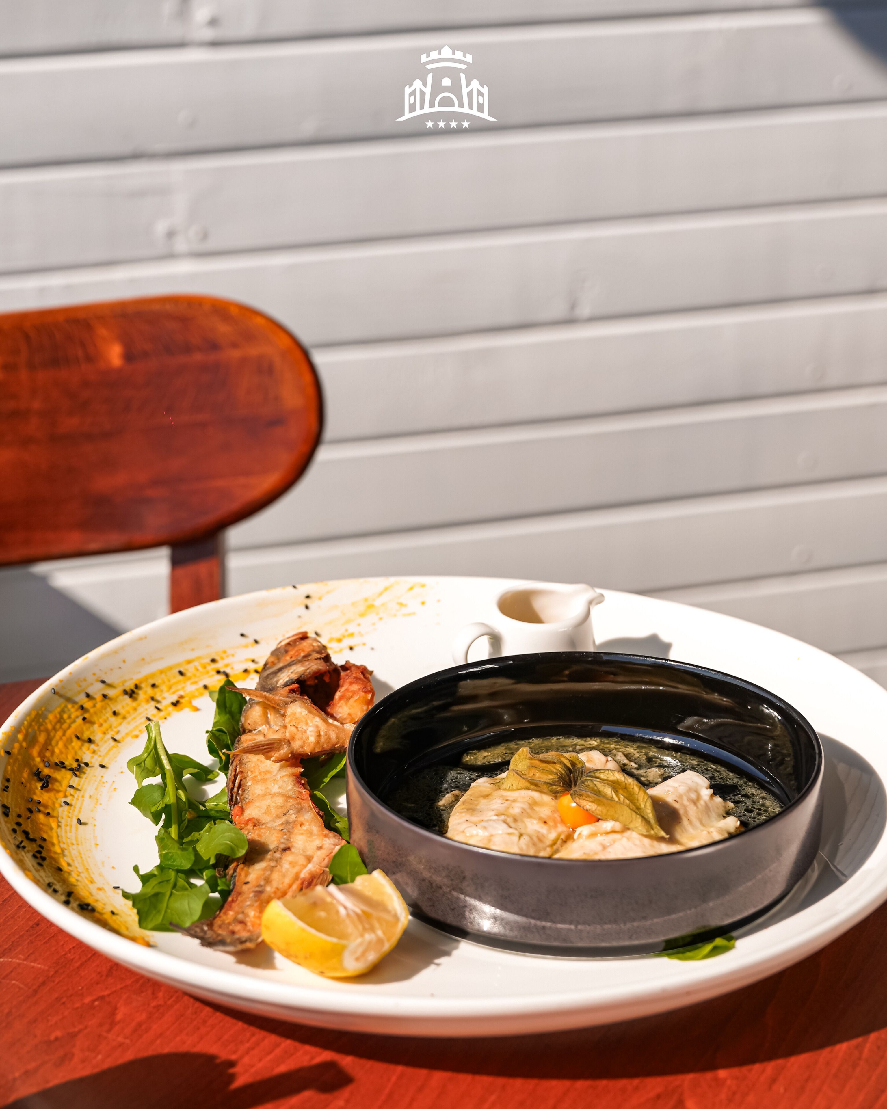 A plate with grilled fish, lemon, and greens, and a black pot with a lid on a table.