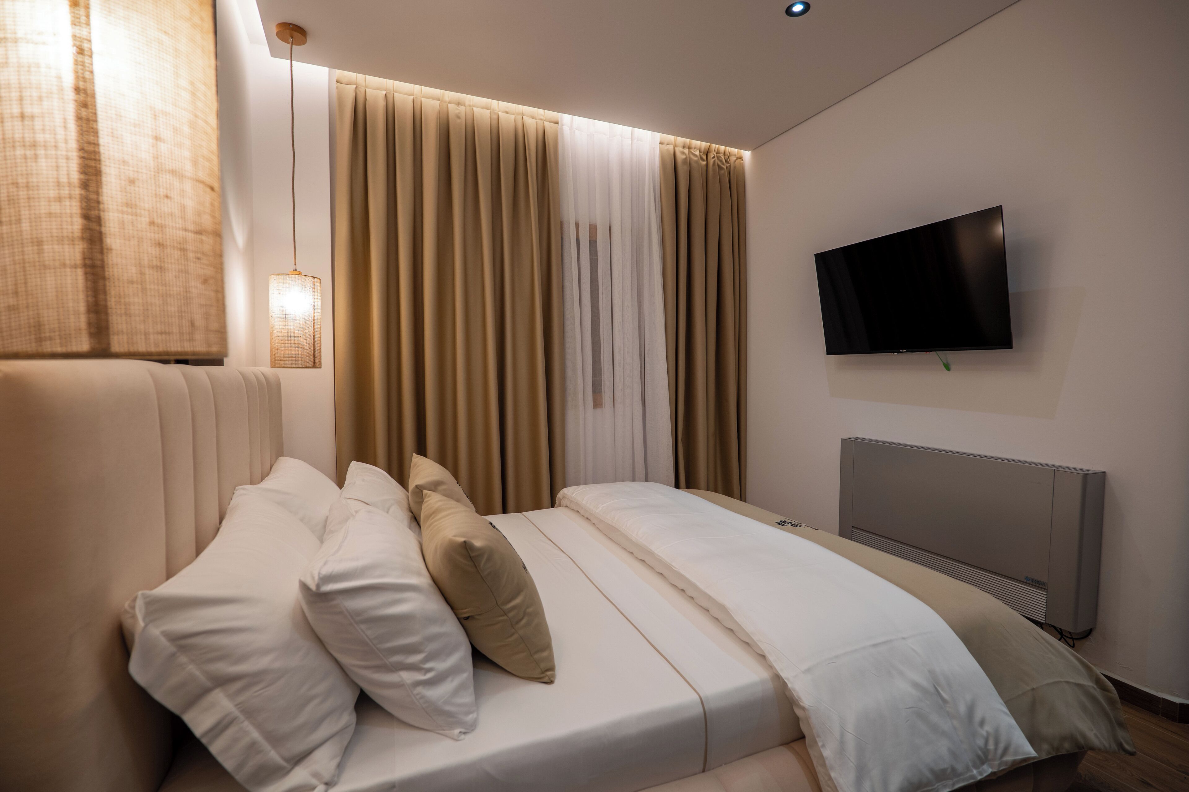 A modern bedroom with a large bed, beige curtains, a mounted TV, and a wall-mounted heater.