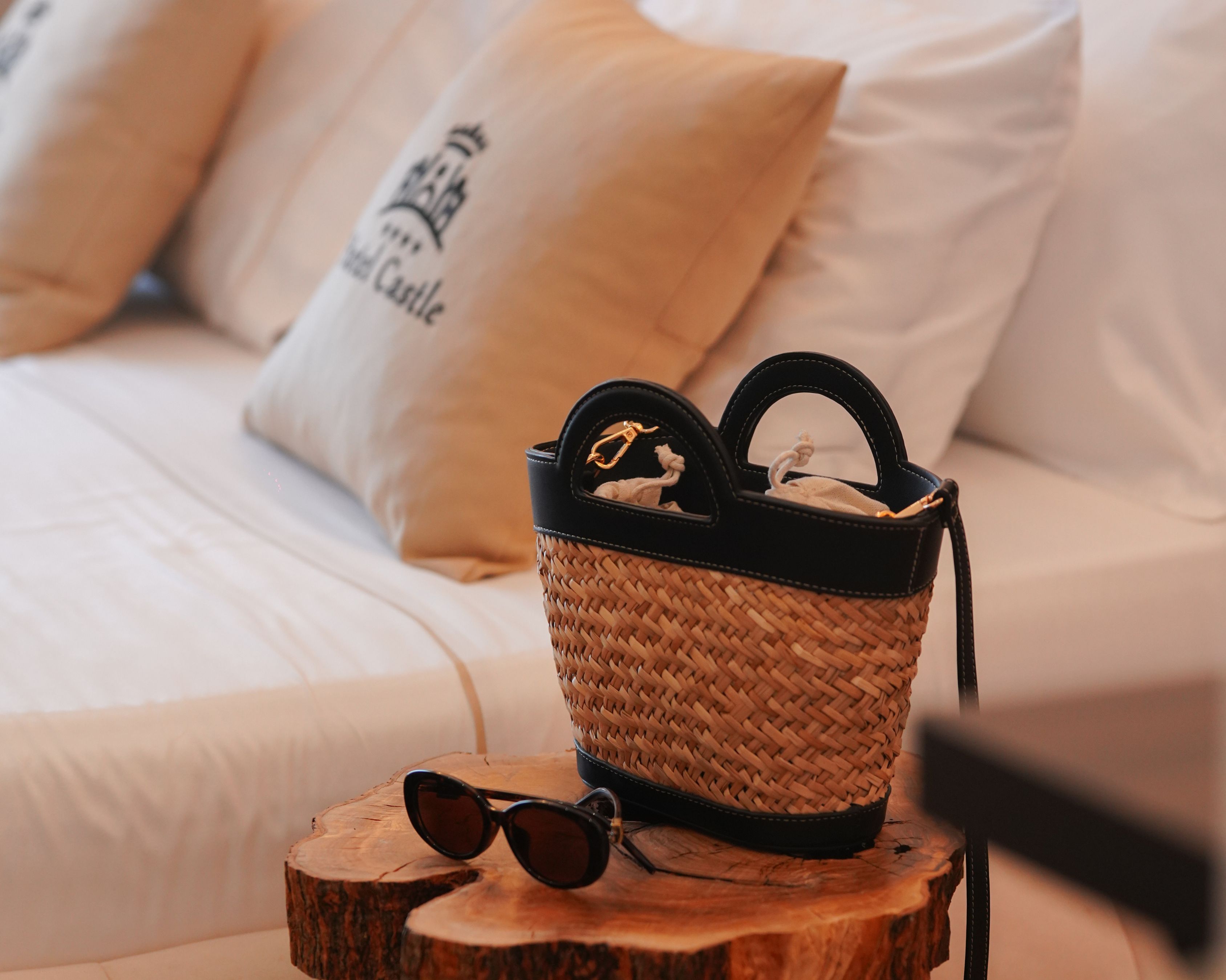 A wicker handbag on a wooden stool with sunglasses, placed on a bed with white bedding and a pillow with text.