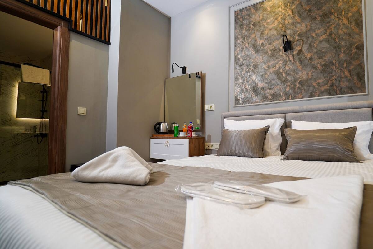 A neatly made bed with pillows, a bedside table with a lamp, a mirror, and a wall with a textured artwork.