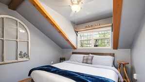 Une chambre avec un grand lit, une tête de lit en bois, une fenêtre avec vue, un ventilateur de plafond et un petit tabouret en bois.
