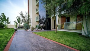 A pathway leading to a modern building with a glass facade, surrounded by well-maintained grass and various plants. - Alexander Palace Hotel Apartment (Amrahia)