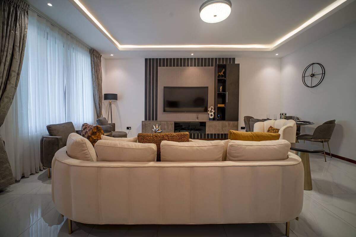 A modern living room with a large beige sofa, a flat-screen TV mounted on the wall, a round coffee table, and a dining area with chairs.