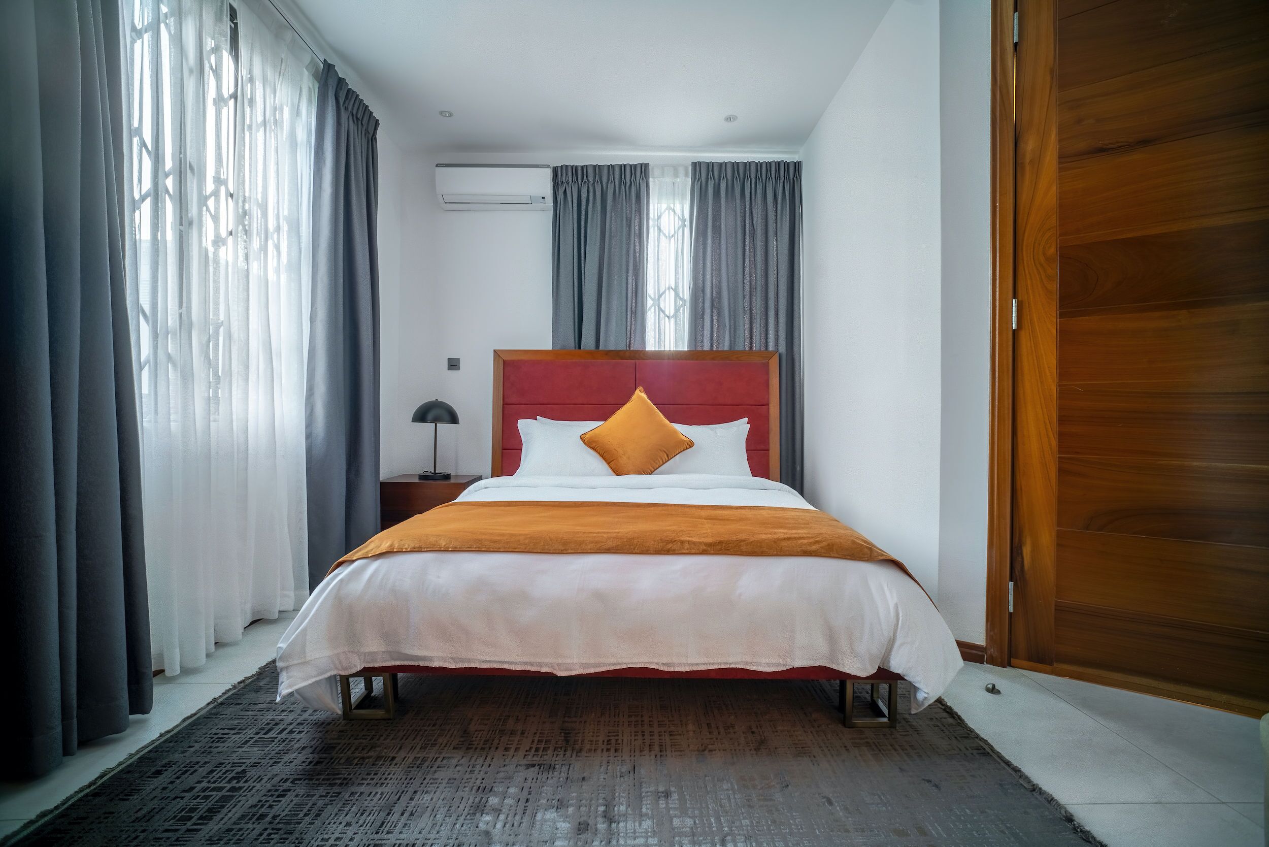 A neatly made bed with a red headboard, a grey curtain, and a wooden door.