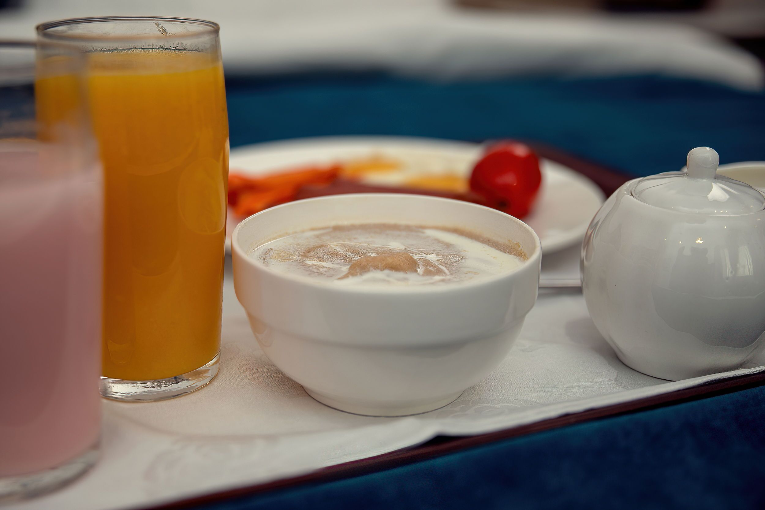 A tray with a glass of orange juice, a bowl of cereal, a small plate with a cherry tomato, and a teapot.