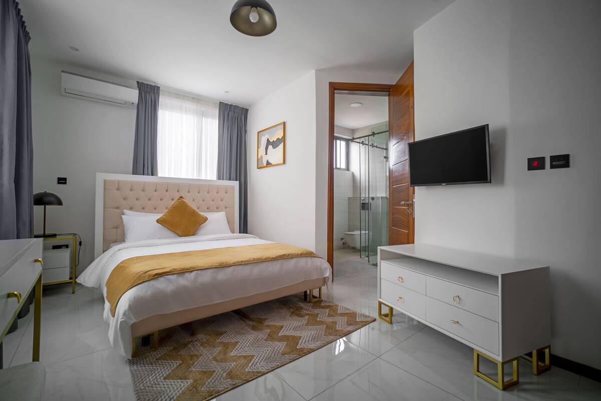 A modern hotel room with a large bed, a television, a dresser, and a bathroom visible through an open door.