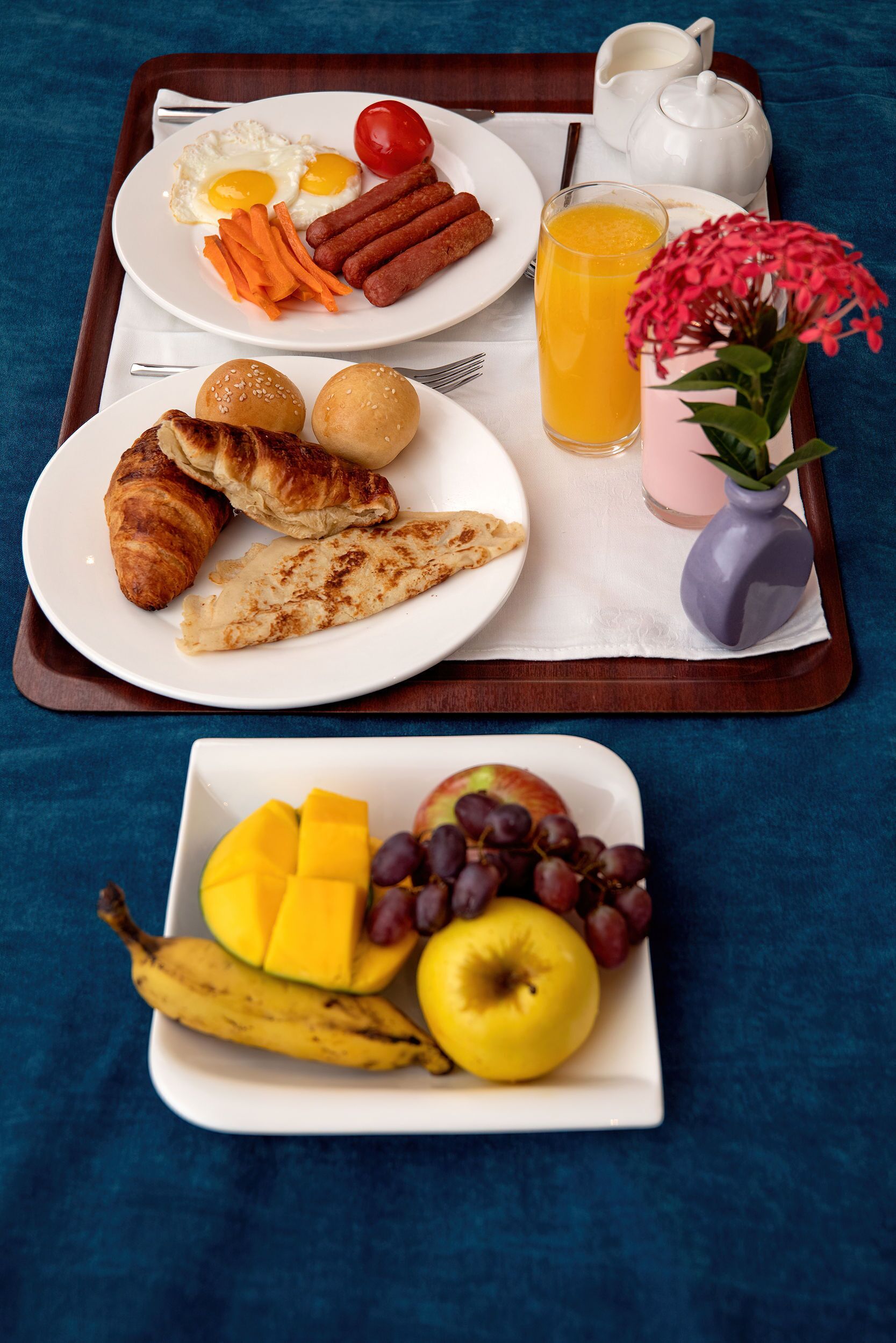 A breakfast tray with croissants, eggs, sausages, tomatoes, carrots, and a glass of orange juice.