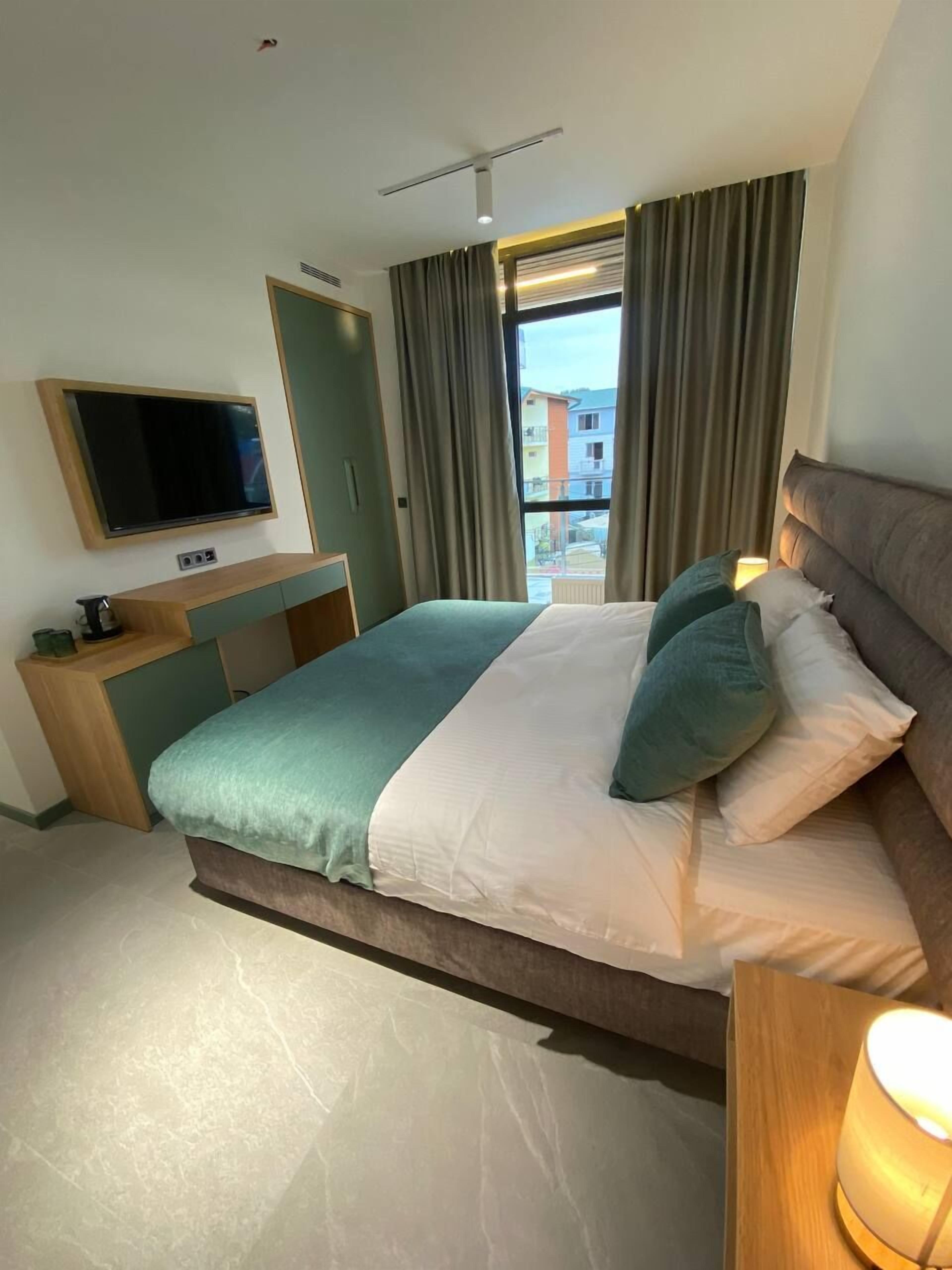 A modern hotel room with a large bed, a flat-screen TV mounted on the wall, a bedside table with a lamp, and a view of buildings through the window.
