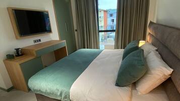 A modern hotel room with a large bed, a flat-screen TV mounted on the wall, a bedside table with a lamp, and a view of buildings through the window.