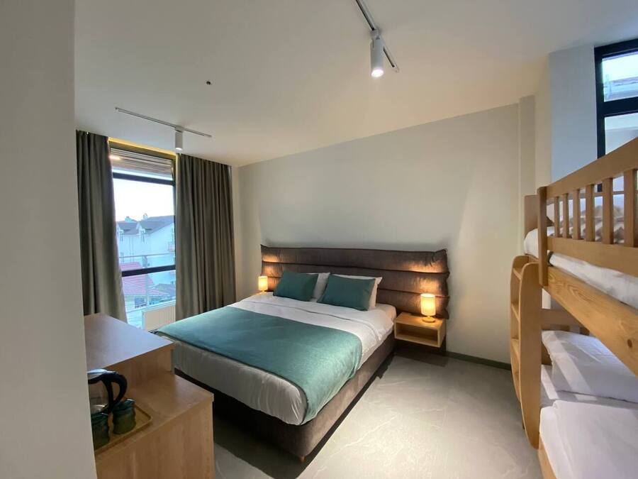 Een moderne hotelkamer met een groot bed, bedlampen, een houten stapelbed, een bureau met waterkoker en een raam met gordijnen.