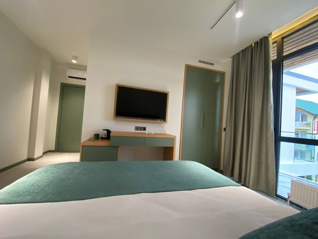 A hotel room with a bed, a TV mounted on the wall, a desk, and a window with curtains.