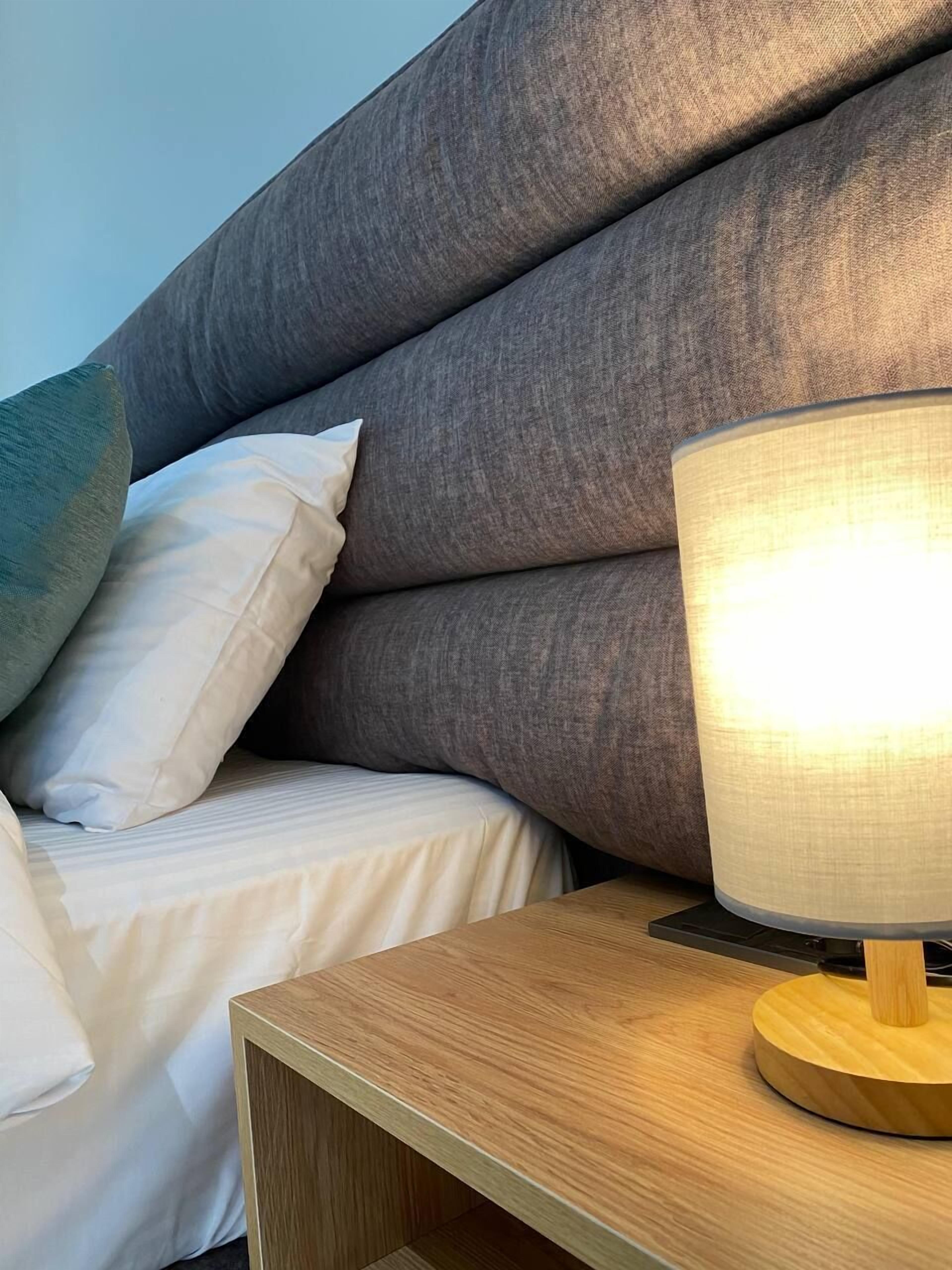 A neatly made bed with a grey upholstered headboard and white bedding. A wooden bedside table with a lamp is visible.
