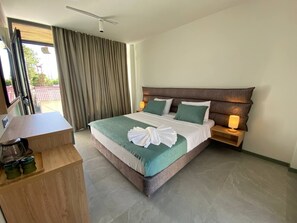 Un cuarto de hotel con una cama grande, un escritorio de madera, una mesita de noche con lámpara y vistas al exterior a través de una ventana con cortinas.