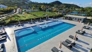 A swimming pool with lounge chairs and umbrellas, surrounded by a landscaped area with a view of hills and houses. - Luxury Waterfront Villa 431F North Beach (Jolly Harbour)