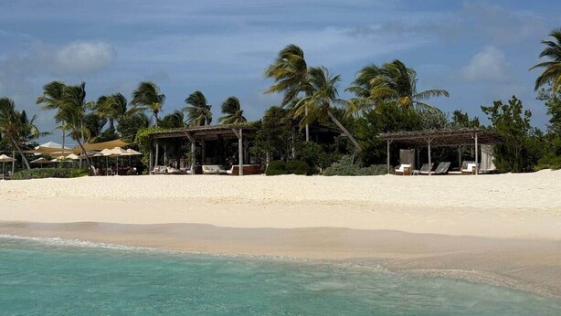 A beach with white sand, clear turquoise water, and palm trees. There are structures with umbrellas and lounge chairs. - Luxury Waterfront Villa 431F North Beach (Jolly Harbour)