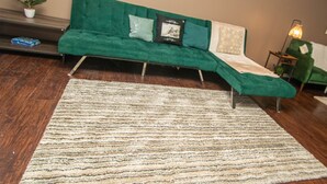 Un canapé vert, surmonté d’un tapis à motifs, dans une pièce dont le sol est recouvert de planches de bois.