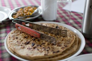 A plate of flatbreads with a utensil on it, a glass of water, and a metal container in the background. - COVIE Bannerghatta 141 (Bengaluru)