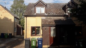 Il s’agit d’un immeuble résidentiel avec un toit brun, une petite fenêtre de grenier et une porte d’entrée. On peut voir trois poubelles devant l’immeuble, chacune étant munie d’une étiquette indiquant un numéro.